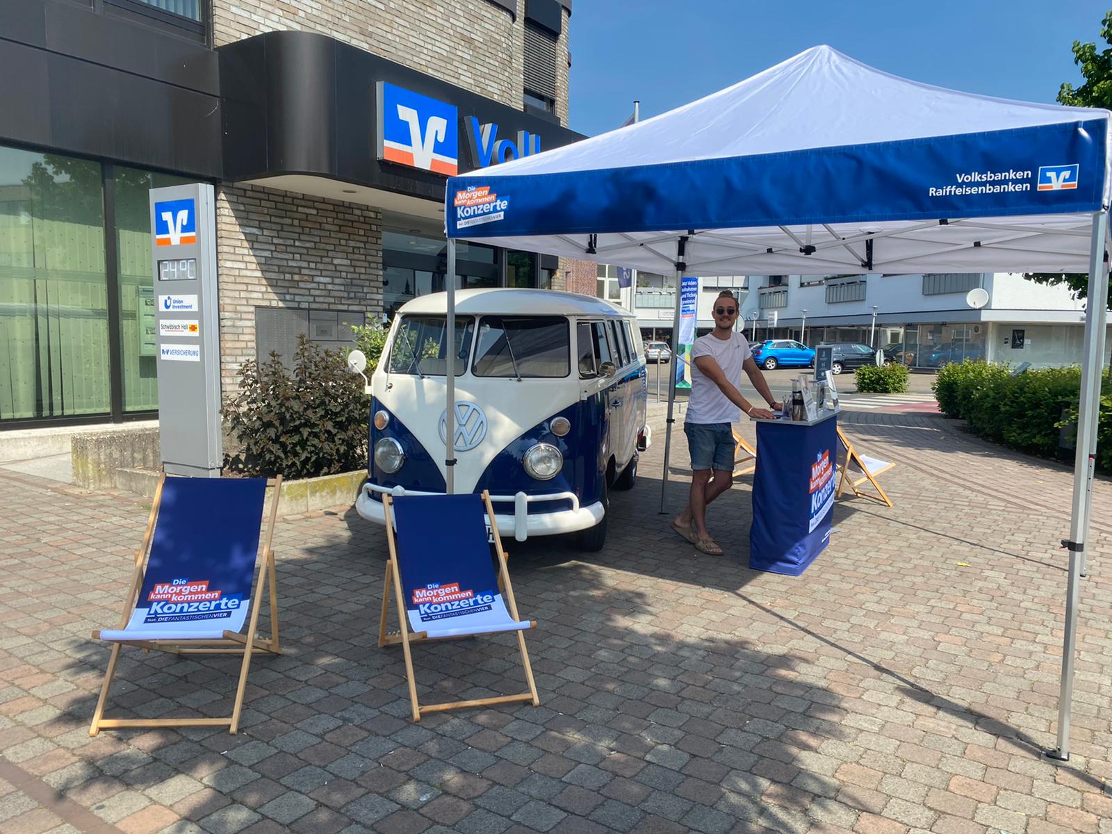 Ein Mann steht an einem Tisch unter einem blauen Zelt neben einem Oldtimer-Volkswagen mit zwei gebrandeten Liegestühlen vor einer Volksbank-Filiale.