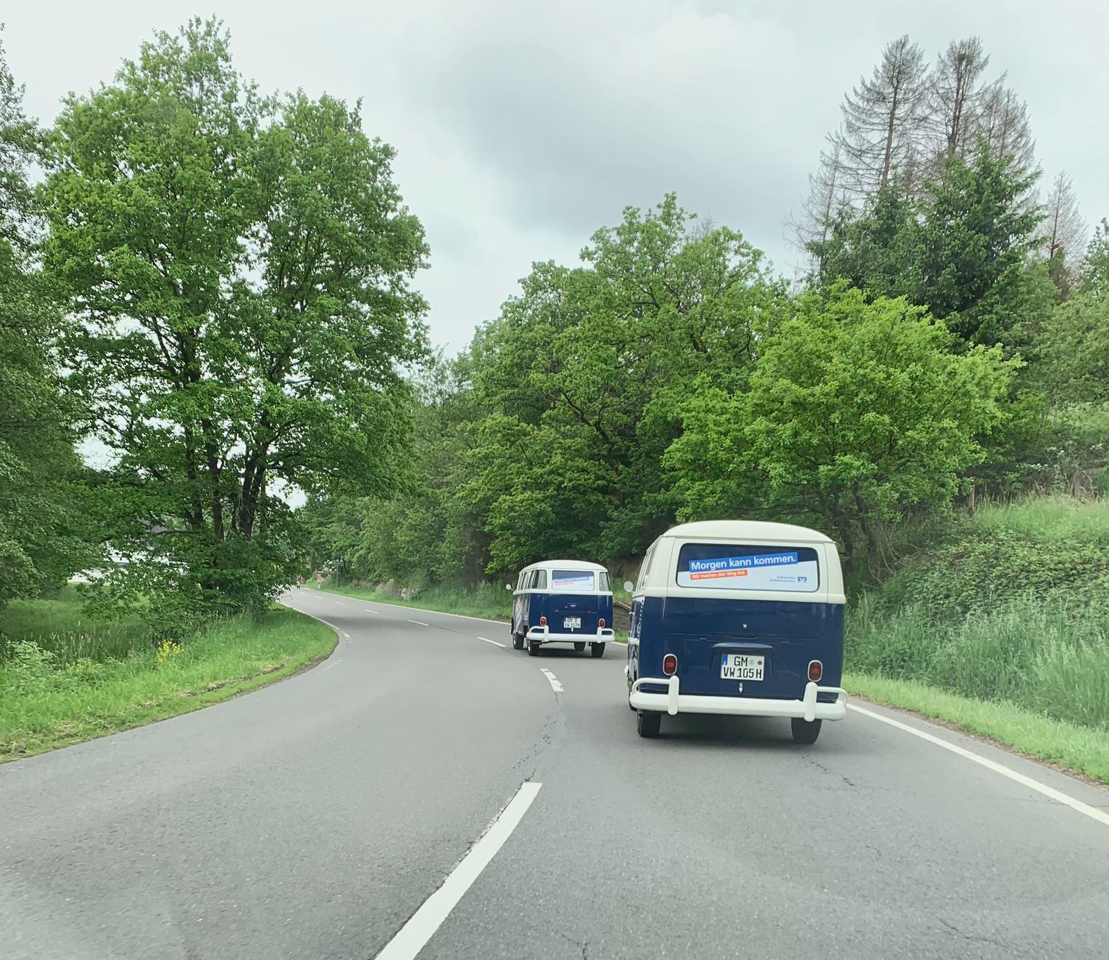Zwei blaue VW-Bullis mit „Morgen kann kommen.“-Aufschrift fahren auf einer Landstraße durch grüne Waldlandschaft.