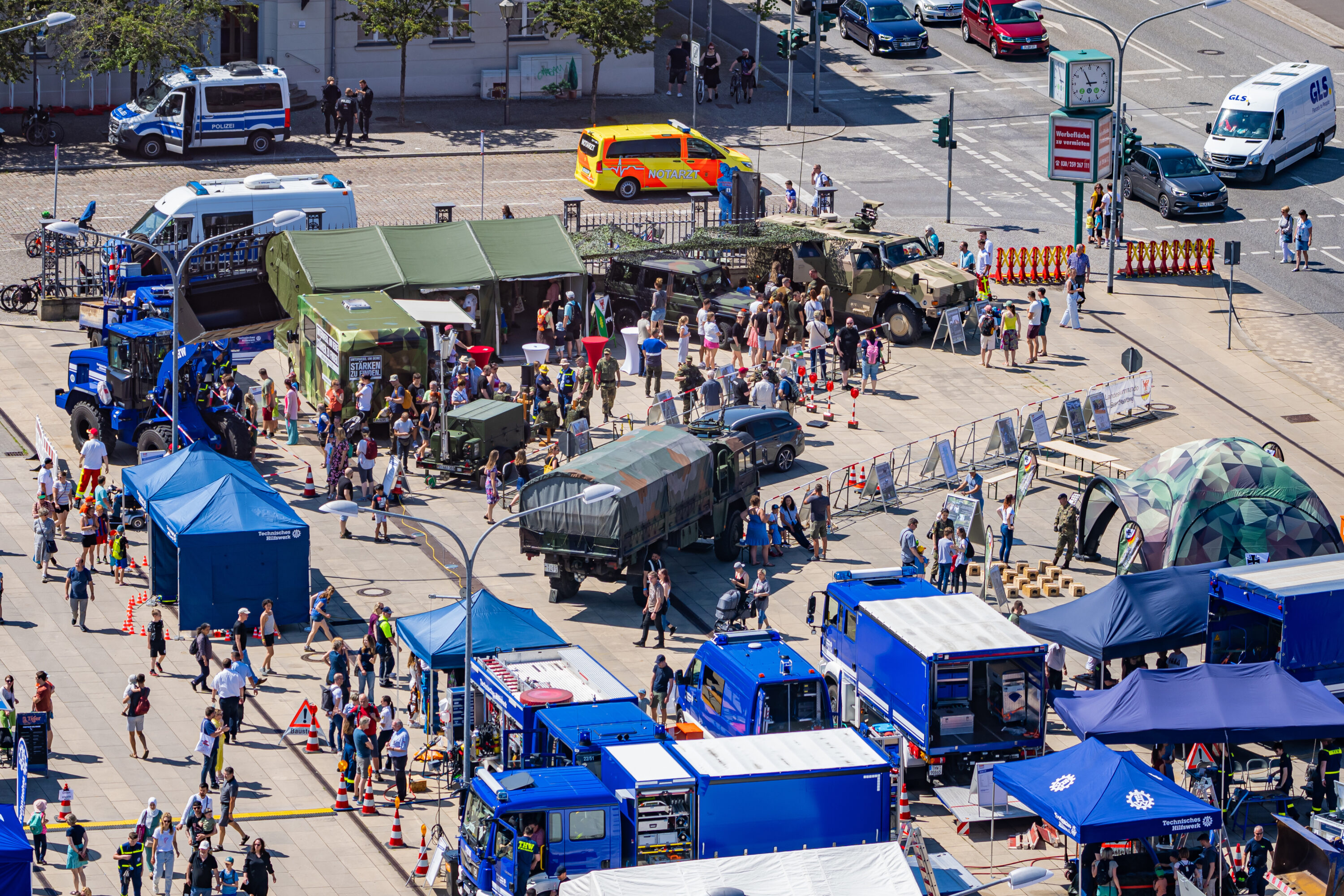 Luftaufnahme eines belebten Veranstaltungsbereichs mit zahlreichen Infoständen, Fahrzeugen von Bundeswehr und THW sowie vielen Besucher*innen. Im Bild zu sehen sind unter anderem Militärfahrzeuge, ein getarnter Pavillon und mehrere blaue Einsatzfahrzeuge.