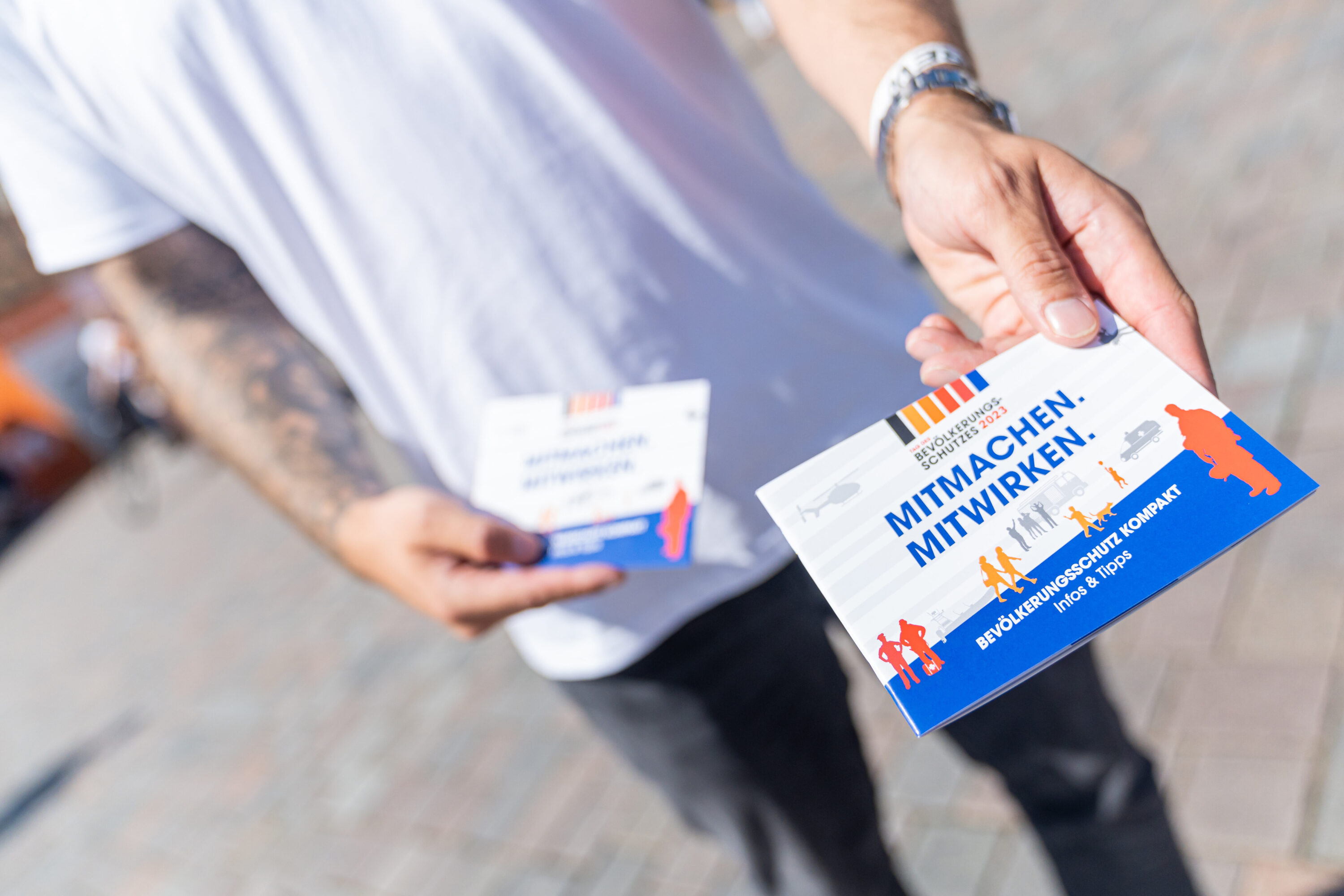 Eine tätowierte Person in weißem T-Shirt hält einen Flyer mit dem Titel „MITMACHEN. MITWIRKEN.“ in die Kamera – es handelt sich um Informationsmaterial zum Bevölkerungsschutz Kompakt, das Tipps und Hinweise zur aktiven Teilnahme vermittelt.