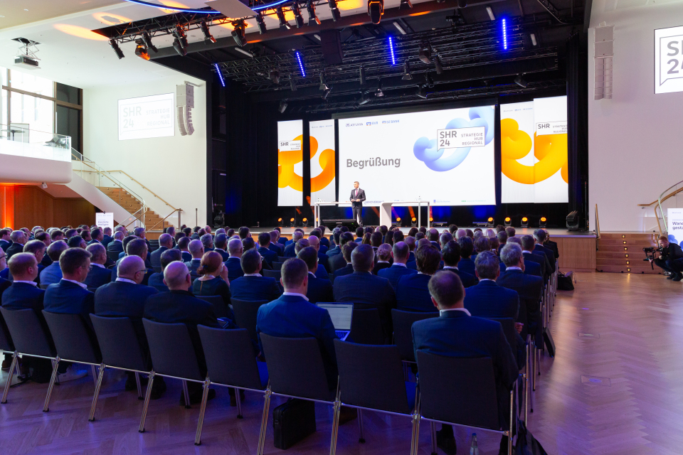 Großer Konferenzsaal mit Publikum, das einer Begrüßungsrede auf der SHR 24-Bühne zuhört.