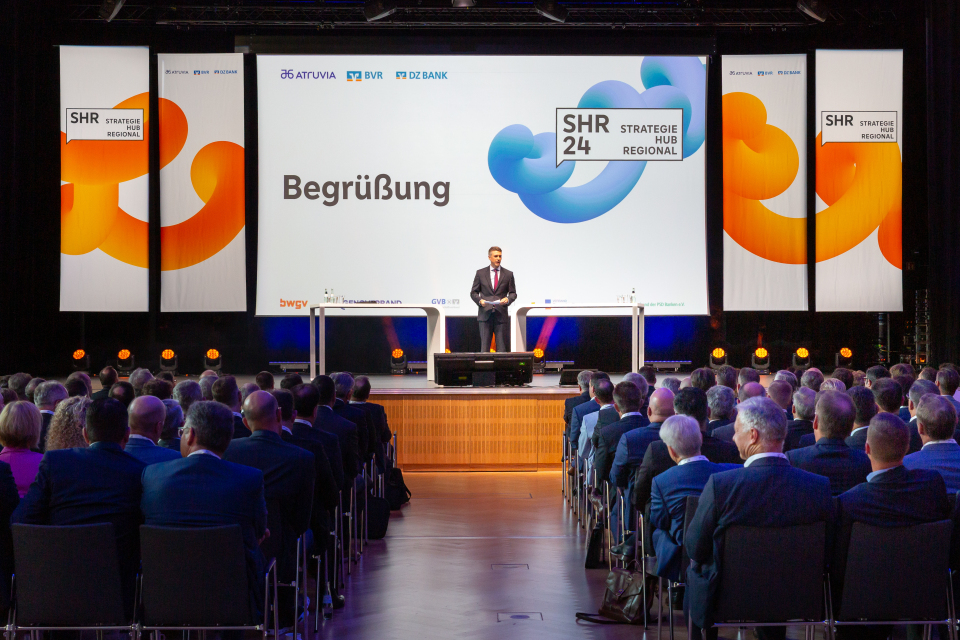 Ein Redner steht auf einer Bühne vor einem großen Publikum in Businesskleidung beim Strategie Hub Regional (SHR 24); auf der Leinwand hinter ihm ist „Begrüßung“ zu lesen, daneben die Logos von Atruvia, BVR, DZ BANK und weiteren Partnern. Die Bühne ist modern beleuchtet, flankiert von Bannern mit dem SHR-Logo und geschwungenen Farbgrafiken in Orange und Blau.
