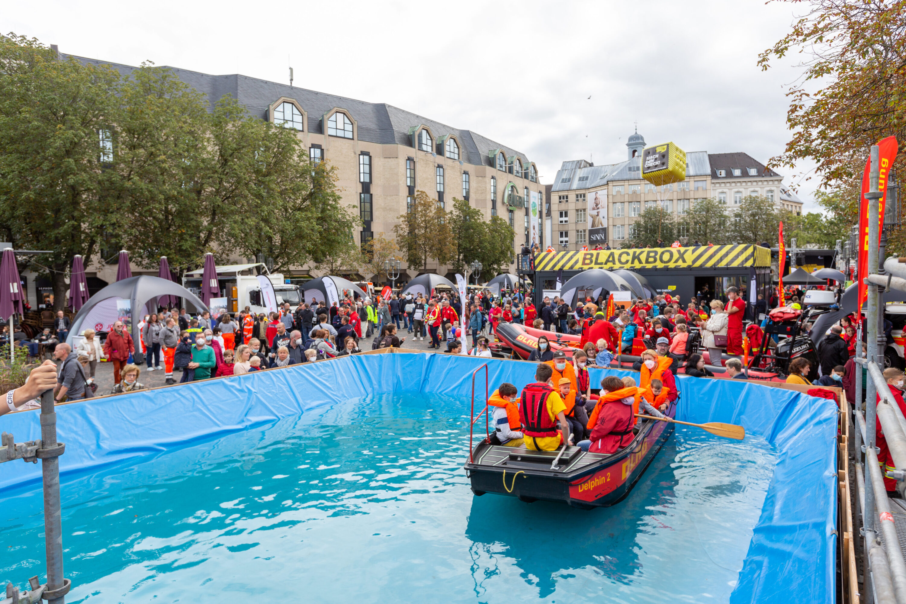 Großes Becken mit einem Rettungsboot der DLRG, besetzt mit Erwachsenen und Kindern in Schwimmwesten – umgeben von vielen Zuschauer*innen beim Katastrophenschutztag NRW 2021 in Bonn. Im Hintergrund: Infozelte, Einsatzfahrzeuge und die markante „BLACKBOX“-Station.