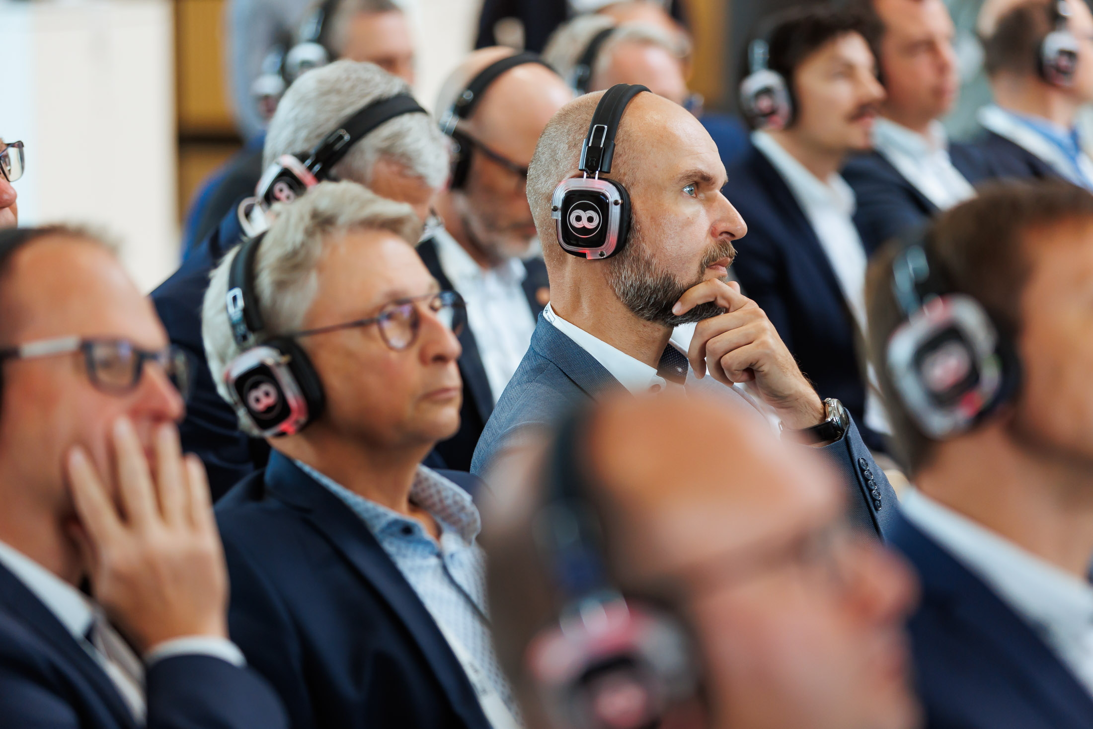 Teilnehmende einer Konferenz verfolgen konzentriert eine Präsentation mit Funkkopfhörern im Silent-Conference-Format; die Männer in Businesskleidung sitzen dicht beieinander und wirken aufmerksam und nachdenklich.