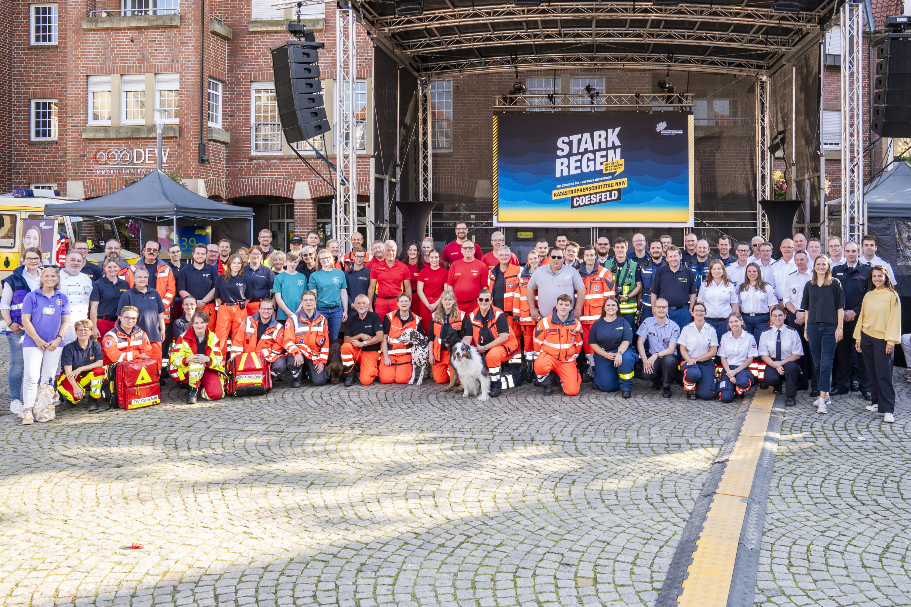 Gruppenfoto von Einsatzkräften verschiedener Hilfsorganisationen vor einer Bühne beim Katastrophenschutztag NRW in Coesfeld. Im Hintergrund ein großes Display mit dem Motto „Starkregen – Katastrophenschutztag NRW Coesfeld“.
