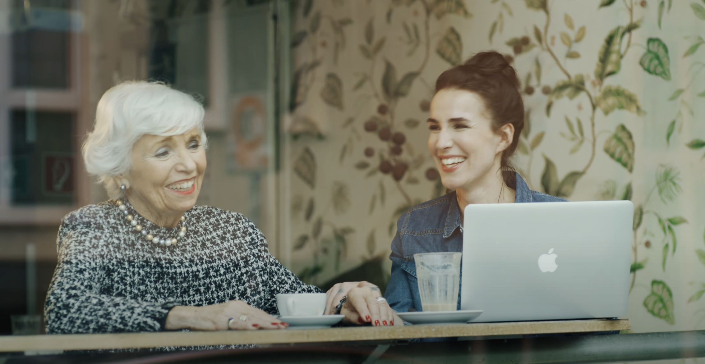 Zwei Frauen unterschiedlicher Generationen sitzen lachend an einem Café-Tisch, eine mit Laptop, die andere mit Kaffeetasse. Die Szene wirkt warm und vertraut – ein Moment digitaler Verbundenheit und echter Nähe.